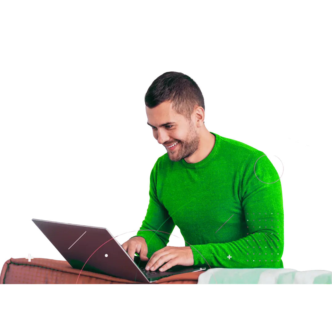 A smiling young man in a bright green long-sleeve shirt happily working on a laptop.
