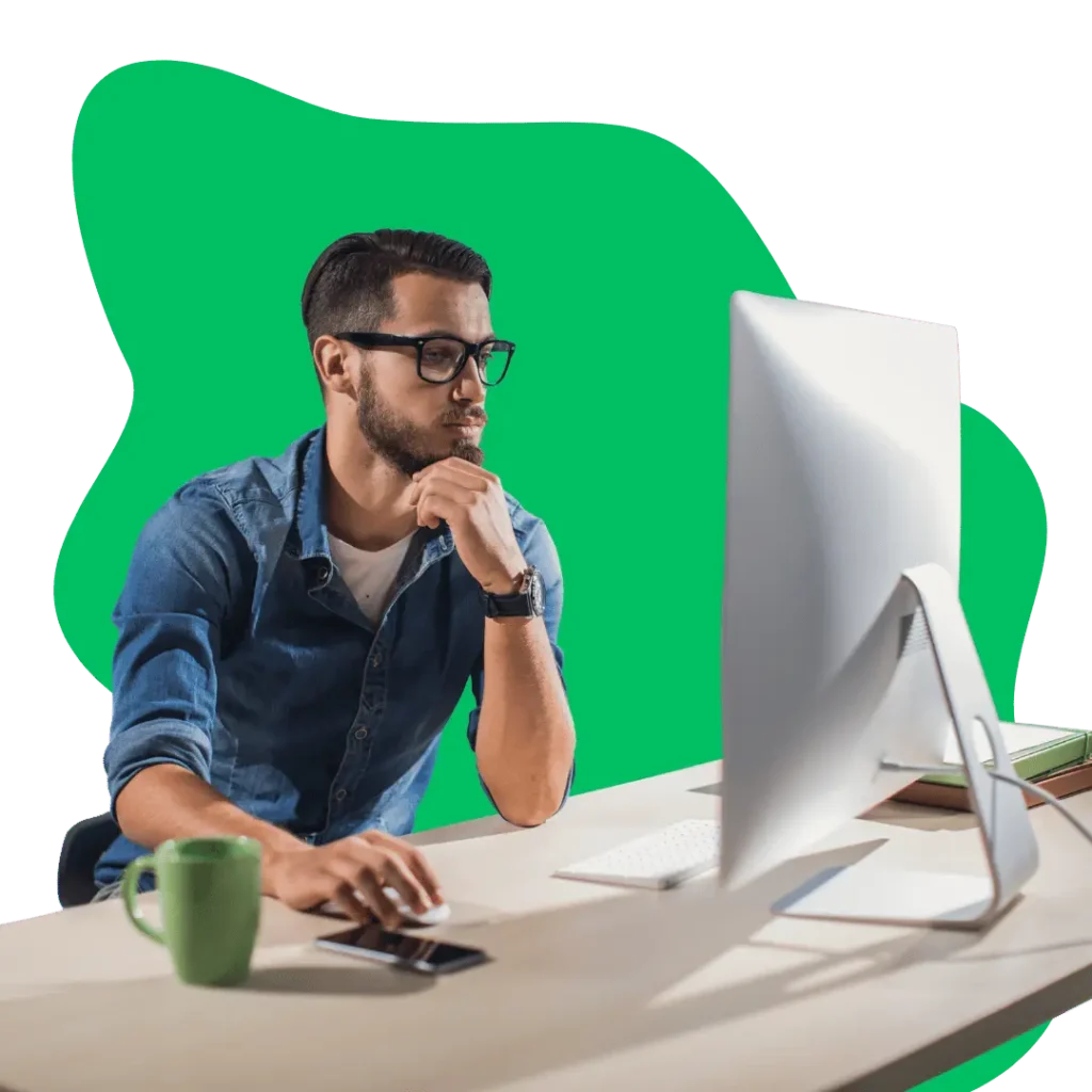A focused man in glasses and a denim shirt intently working on a desktop computer, with a thoughtful expression