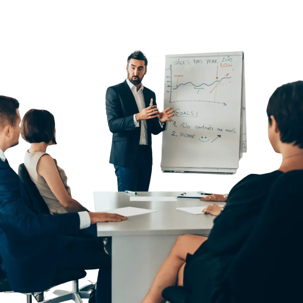 A male presenter in a suit gestures towards a flip chart with sales goals and growth charts, addressing three people at a business meeting.