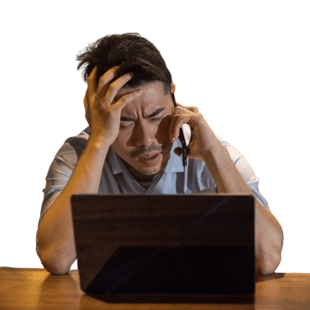 A man appears frustrated and stressed, holding a phone to his ear with one hand while the other is on his head, looking down at an open laptop.