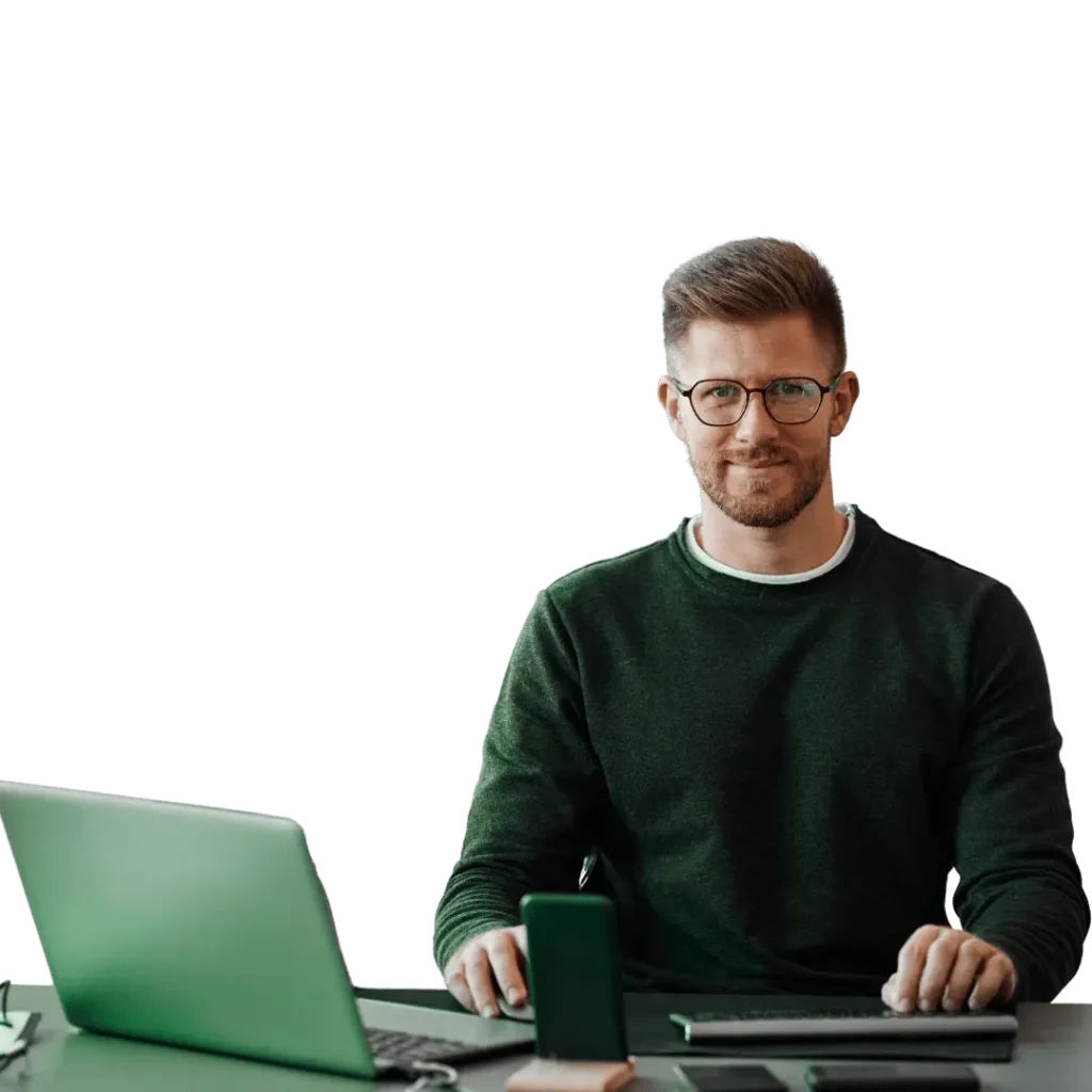 A focused male professional wearing glasses and a dark green sweater, working at a desk with a laptop, smartphone, and tablet