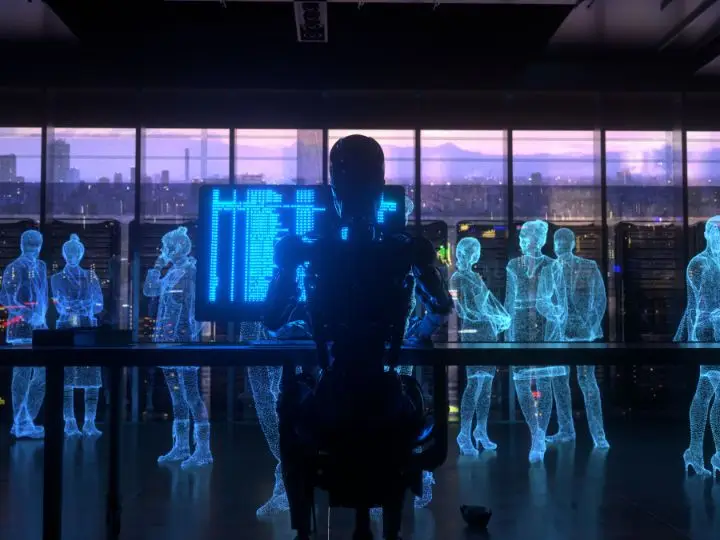 A humanoid robot sits at a desk in front of a glowing digital screen, surrounded by holographic figures of people, inside a futuristic office